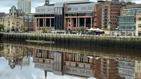 An imposing court building made from light red stone with large dark windows and a silver roof reflected in a river running in front of it.