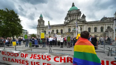 Getty Images A man in a rainbow flag watches a Christian protest against the Belfast Pride parade