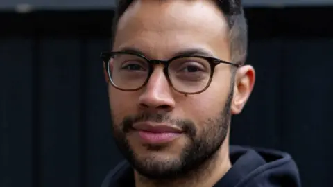 Lanre Bakare Headshot of Lanre, who looks into the camera wearing black and brown rimmed glasses and a black hoodie.