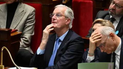 Michel Barnier wearing a blue shirt and tie and black suit sits a session of the National Assembly