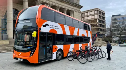 An orange and white bus parked outside Sheffield's City Hall. The bus has the words South Yorkshire People's Network written on the side. In front of the bus a man stands looking at a row of five black and orange e-bikes
