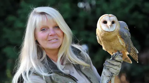A long grey-haired woman has an owl perched on a gauntlet which is on her left hand.