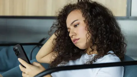 A young woman with a confused facial expression sits on a sofa, looking at her smartphone.