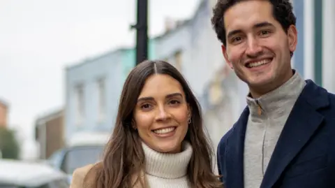 PA Close up of couple smiling. She is wearing a white jumper and he is wearing a grey top. They are stood in front of houses which are blurred in the background.