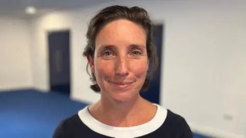 A woman with dark brown hair and blue eyes smiles at the camera, wearing a black top with a white neckline. In the background is a blue floor with white walls.