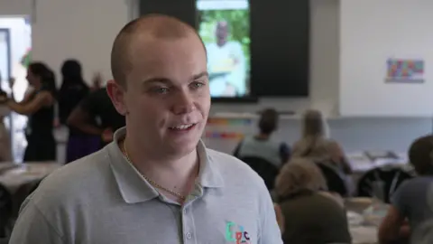 Bradley Oliver, who is 23. He has very short light brown hair. He is wearing a grey jersey polo top and gold necklace. He is smiling and behind people are sitting at tables in the Deaf café signing to each other.