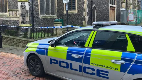 A marked fluorescent yellow and blue police car is parked near a church which is near an alleyway. In the background there is white and blue police tape used to cordon off an area of the street.