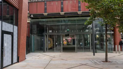 The entrance to Salford Central, which features glass doors set below a redbrick building. The name of the train station and a rail symbol can be seen in silver lettering across the entranceway. 