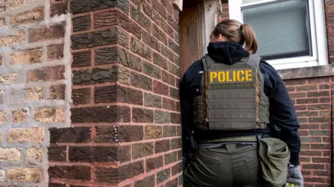 A woman with a vest labelled "police" appears poised to enter a brick house while conducting an immigration raid