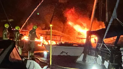 Bright orange flames coming off a boat, with firefighters close to the scene
