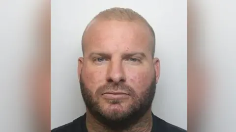 Mugshot of Alex Male in a black t-shirt. He has short blond hair, a light brown beard and a neck tattoo