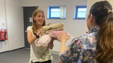 A woman holding a number of items that look like blankets, with a woman handing them to her. They both have long hair. 
