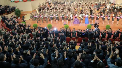 KCNA North Korean leader Kim Jong-un, centre, waves during a celebration, surrounded by high-ranking military officials and nuclear scientists. In the background, an orchestra and row of standing women can be seen on stage