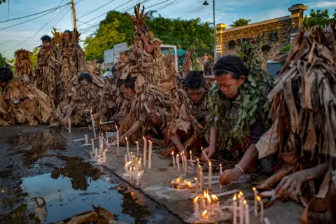 Ezra Acayan/Getty Images Devotees covered in mud and dried banana leaves light candles and pray outside a closed church