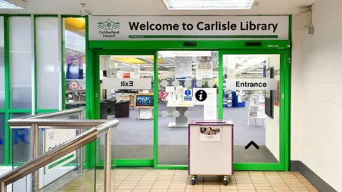 Front facade of Carlisle Library. There are glass doors with green panels and signage reading 'Cumberland Council'. There is a book donation box outside. 