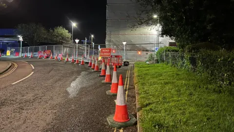 A long line of traffic cones have been placed down one side of a road. Metal barriers are seperating the road from a building. A road closed sign is on display. Street lights are illuminated as it is dark.