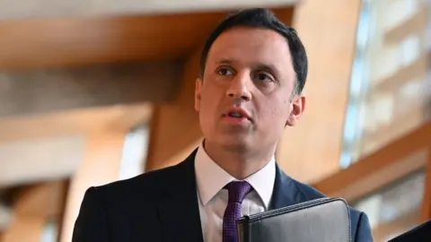 Getty Images A man with dark hair walks in the Scottish Parliament. He is holding a black folder tucked up against his chest. He is wearing a dark suit, white shirt and purple tie. 