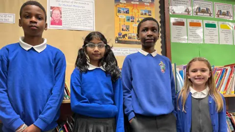 Four primary school pupils - two boys and two girls - smile at the camera. They are wearing uniforms with blue sweaters and stand in front of a colourful library noticeboard.