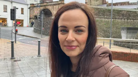 Shows a woman with dark hair and a brown jacket with the Derry Walls and Ferryquay Gate in the background.