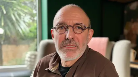 Simon Bessell is wearing round black glasses and a brown sweatshirt. He has a grey beard.