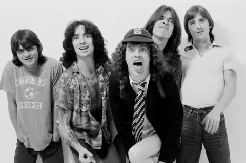 Getty Images AC/DC IN 1979 - from L-R Malcolm Young, Bon Scott, Angus Young, Cliff Williams and Phil Rudd. The group pose in a black and white promotional photo, all looking into the camera and smiling or making faces.