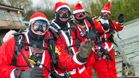 Vobster Quay Four scuba divers dressed in Santa outfits waving at the camera at the water's edge