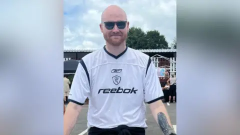 Sam Bradley-Green Sam Bradley-Green has a shaved head, brown stubble, sunglasses, a tattoo on his inner left arm and wears a white short-sleeved Bolton Wanderers top.