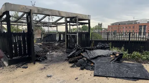A wooden structure has been turned completely black after a fire. There us debris out in front of the structure, which appear to be charred panelling. The structure now has now roof with just three or four thin beams drooping across the empty void in the middle. 