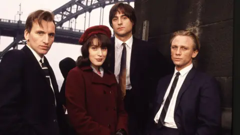 A photo from the set of Our Friends in the North which features a young Christopher Eccleston on the left who is in a black suit and tie with brown floppy hair and is looking into the camera with a serious look on his face. Gina McKee is standing beside him and is wearing a deep red jacket and hat. She has shoulder-length dark hair and is looking into the camera with a slight smile. Mark Strong is centre-right and is standing slightly higher up than the others. He has long, brown hair and is wearing a dark suit with a brown tie and is looking into the camera with a straight face. Daniel Craig is on the right and is leaning against a stone pillar. He has his blonde hair swept back and is wearing a dark suit. Part of the Tyne Bridge is in the background above them.