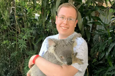 Handout A man with mousy brown short hair pushed away from his forehead is wearing rectangular glasses, smiling at the camera and holding a koala which is also looking at the camera and has one of its paws resting on his arm. He is wearing a white top and has his hand around the koala with trees in the background.