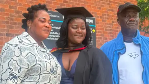 A woman in the middle is wearing a black graduation gown. Another woman on the left is wearing a dark and white top, while a man on the right is in a dark baseball cap and blue and white clothing.