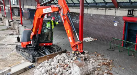 Swindon Borough Council Image of a man in a high vis jacket and a hard hat, waving from a digger in the background can be seen a dilapidated building, covered in graffiti, with rubble and large sections of concrete in the foreground. 