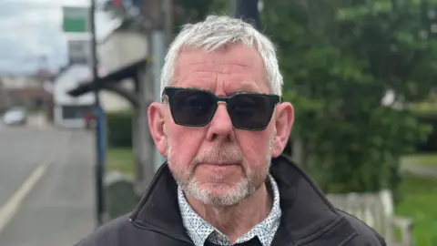 George King/BBC A head and shoulders image of Bill Bulstrode. He is wearing a black jacket over a shirt and dark sunglasses. He is looking directly into the camera. 
