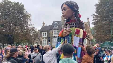 The puppet is surrounded by people taking photos as she makes her way through the park. She is draped in a patchwork blanket.