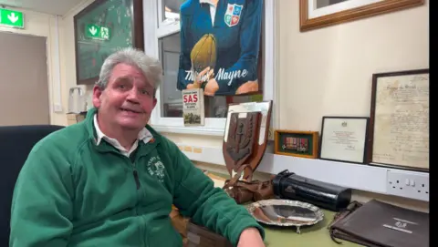 David McCallion, a man with grey hair and a green fleece sits in an office surrounded by Blair Mayne memorabilia. To his left on a green desk sits a silver platter and a wooden plaque with the SAS logo.