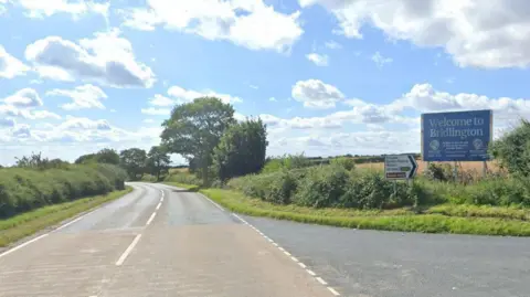A country road flanked by green bushes and trees. There is an exit lane on the right with a big blue sign on the grass verge reading "Welcome to Bridlington". Next to that is a white road sign listing Grindale, Burton Fleming and Malton, with a black arrow pointing right. The sky is blue with scattered white and grey clouds.