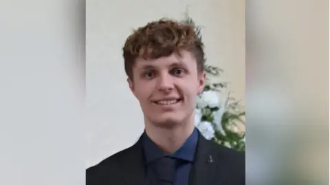 Kacper Dudek is wearing a black suit and tie with a navy shirt. He has brown hair and is smiling into the camera.