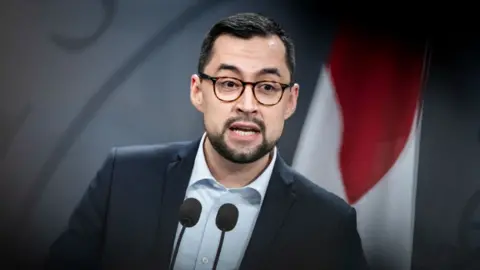 Mute B Egede at a news conference. He has a dark suit jacket on and light blue shirt with no tie. He's wearing tortoise-shell glasses and there's a Greenland flag hanging behind him.