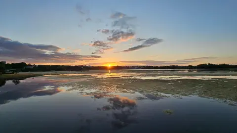 scuddingcloud Clouds and the glint of a setting sun are reflected on a large body of water.