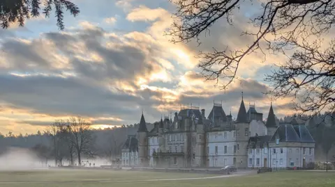 Steven McDonald A large, turreted stone mansion sits in misty morning light. Soft sunlight breaks through dramatic clouds, illuminating the grand building and the trees surrounding the open lawn.