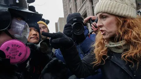 A woman with red hair and wearing a white cable-knit beanie hat points a gloved finger at a man dressed in googles, a hardhat and a pink respirator mask. She looks angry but not afraid. the action is happening in a crowded group of people, a cameraman is taking photographs behind them.