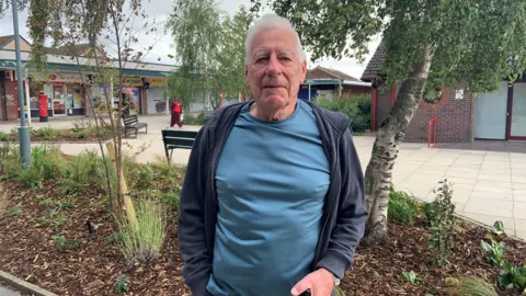 A man with white hair is wearing a blue t-shirt and a navy hoodie. He is standing in a shopping precinct in front of some planting