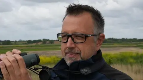 John Fairhall/BBC Aaron Howe holds a pair of binoculars close to his face as he looks out on to marshland behind the camera. He has short dark hair with some grey around the sides. He has glasses. He wears a black jumper.