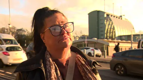 BBC Caroline Willgoose, who has dark hair tied up in a ponytail and black rimmed glasses. She wears a neutral expression while listening to an interviewer's question.