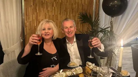 Family handout/PA Stuart Emerson and his wife Claudia celebrating Christmas in 2024. They are sitting at a table and holding their glasses of pink champagne in the air. He is wearing a white shirt and a black blazer, while she is dressed in a black dress with silver bows and a black jacket. On the table there are gold crackers laying on the plates, and the room is decorated with candles and black and gold balloons.