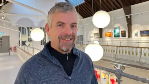 Calum is standing inside a large bright and airy studio space. Behind him are large, globe-like lights.