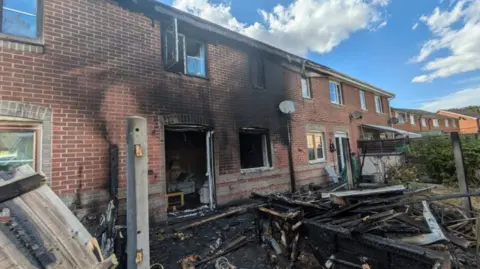 A fire damaged home, with charred wood littering a garden and black soot covering the front of the house. Three windows and doors are open.