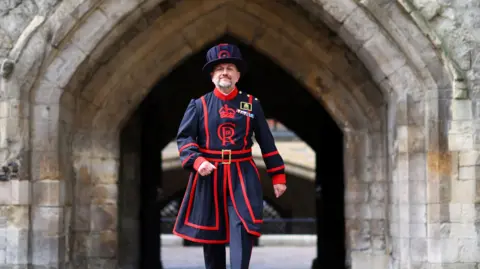 Historic Royal Palaces Andy Harris pictured in the grounds of the Tower of London in the daily blue and red YW uniform. He swings his arms and smiles at the camera.