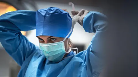 Getty Images A female doctor in a blue gown ties up her face mask, looking into the distance. 