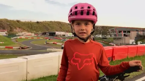 7 year old Rhona Livesey, in an orange cycling top and pink helmet, with the twists and turns of the karting track visible behind her.